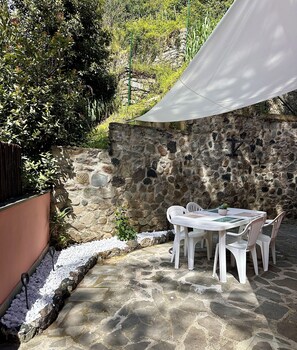 Un patio de piedra con una mesa y sillas blancas, rodeado por un muro de piedra y follaje.
