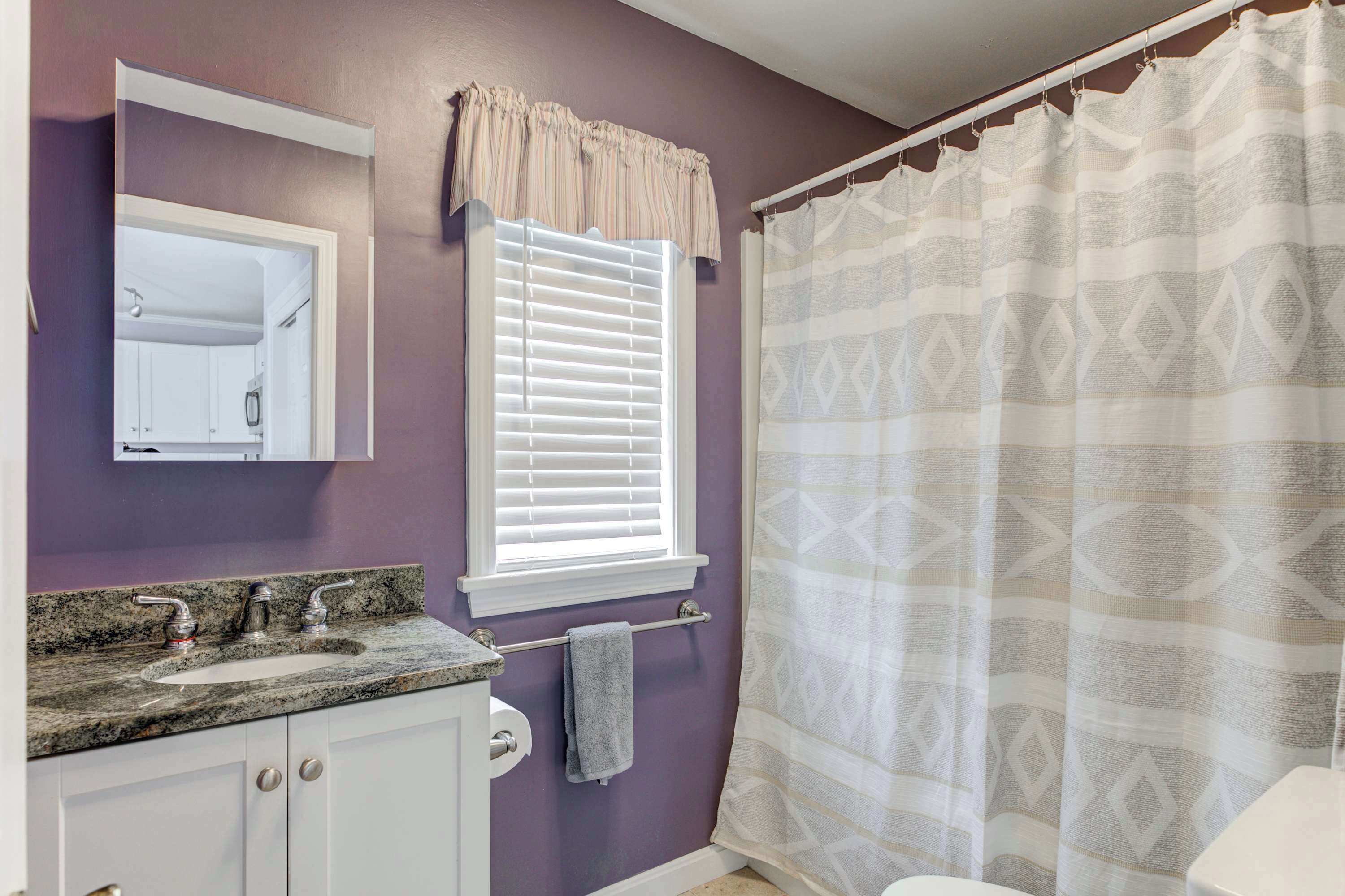 Un baño con una pared morada, un tocador blanco con encimera de granito, una ventana con persianas blancas y una cortina de ducha con diseño geométrico.