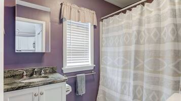Un baño con una pared morada, un tocador blanco con encimera de granito, una ventana con persianas blancas y una cortina de ducha con diseño geométrico.