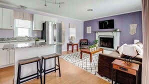 Una cocina moderna con armarios blancos, un refrigerador de acero inoxidable y una isla integrada con taburetes. Hay una chimenea con repisa blanca y un televisor de pantalla plana montado sobre ella.