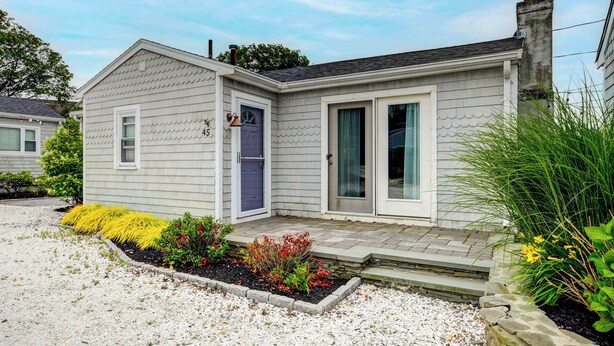 Una casa de un piso con revestimiento gris, una puerta principal con timbre azul y un pequeño jardín delantero con un camino de grava y diversas plantas.