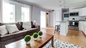 Una sala moderna con un sofá marrón, una mesa de centro de madera y tres plantas en macetas. Se observa una cocina con armarios blancos, un microondas y un horno.