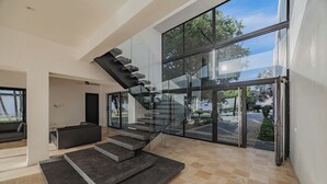 Un intérieur moderne avec un escalier en verre menant à un balcon, un canapé de couleur sombre et un sol carrelé.