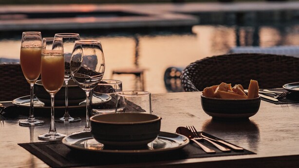 Une table dressée pour un repas, avec des verres remplis d’une boisson, un saladier de fruits et une assiette, donnant sur un coucher de soleil et des palmiers.
