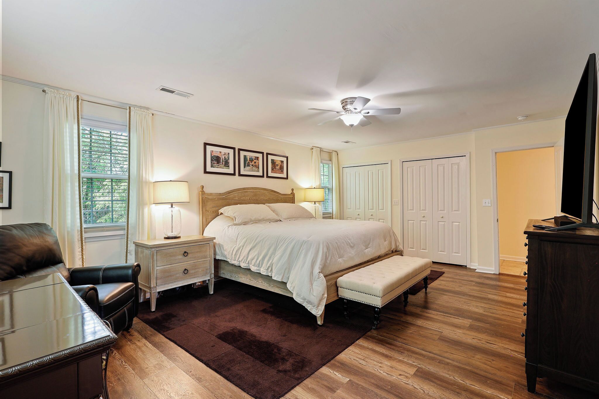 A bedroom with a bed, bedside tables, a bench, a ceiling fan, and a window with curtains.