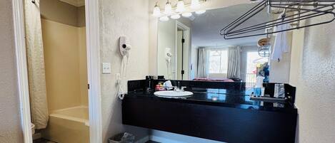 Un baño con encimera negra, lavabo blanco y un espejo con luces. Tiene una cortina de ducha y un perchero.