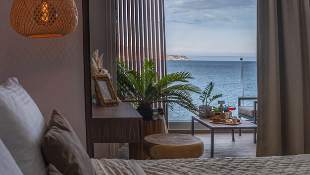 A bedroom with a large bed, a bedside table with a lamp, a view of the sea, and a balcony with a table and chairs. - Coastline of Paradise Luxury (hrakleion)