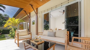 A modern outdoor living area with wooden furniture, a glass table, and a view of a pool and garden. - Villa Emilia with Private Pool (Rawai)