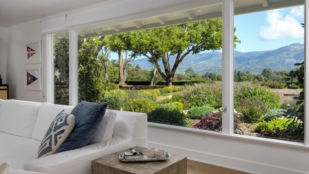 A modern living room with a white sofa, a coffee table, and large windows offering a view of a landscaped garden and mountains. - Stunning Hope Ranch view home, outstanding amenities, private beach. 30 day min. (Santa Barbara)