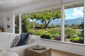 A modern living room with a white sofa, a coffee table, and large windows offering a view of a landscaped garden and mountains.