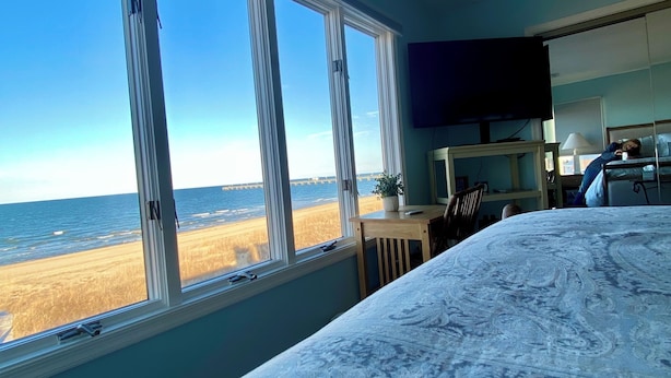 A bedroom with a large window overlooking a beach, a bed with a patterned bedspread, a desk, and a television.