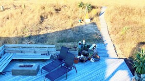 A wooden deck with two lounge chairs and a small table, overlooking a beach and dunes.
