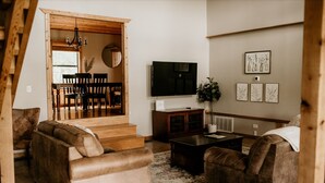 A living room with a sofa, a coffee table, and a TV. There is a staircase leading to a dining area with chairs and a table. - A cozy & charming, German-style home in the heart of the Texas Hill Country (Kerrville)