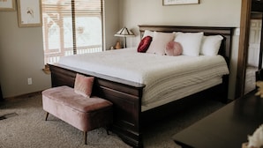 A bedroom with a large bed, a wooden nightstand, a bench, and a window with blinds. - A cozy & charming, German-style home in the heart of the Texas Hill Country (Kerrville)