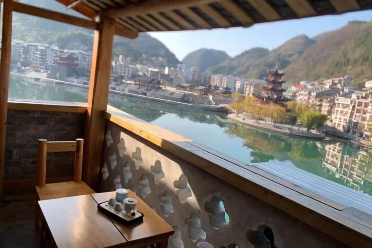 A room with a wooden table, a chair, and a view of a river with traditional buildings and mountains.