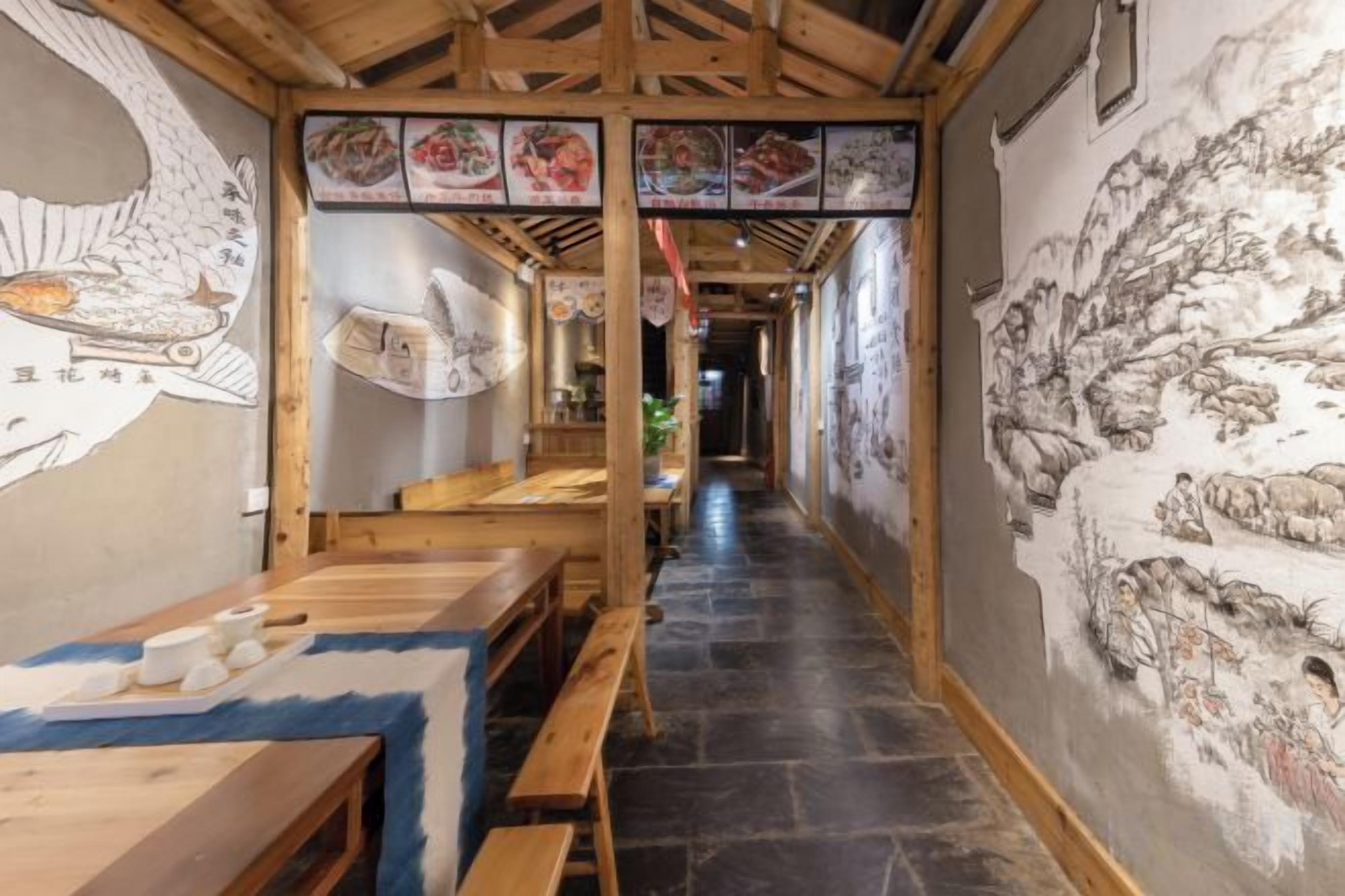 A narrow corridor with wooden benches and tables, traditional wall art, and a tiled floor.