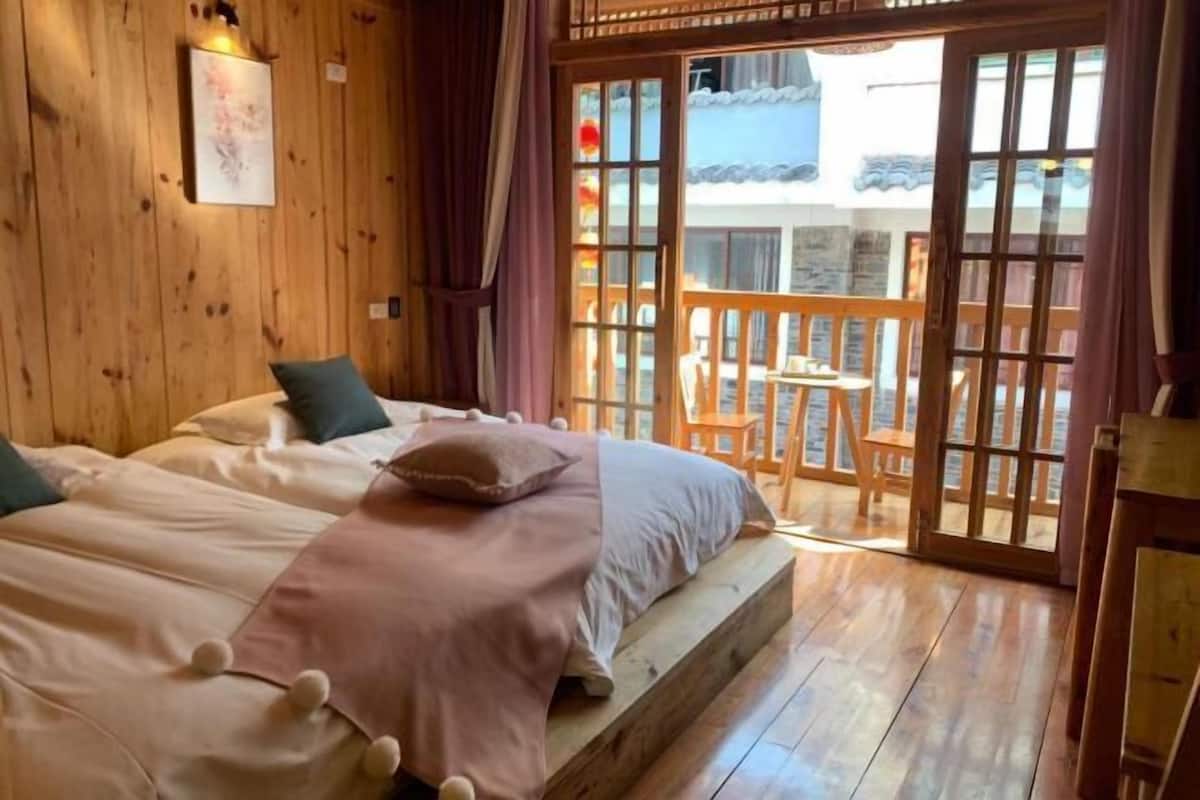 A wooden room with two beds, a balcony with wooden chairs, and a view of a traditional building.