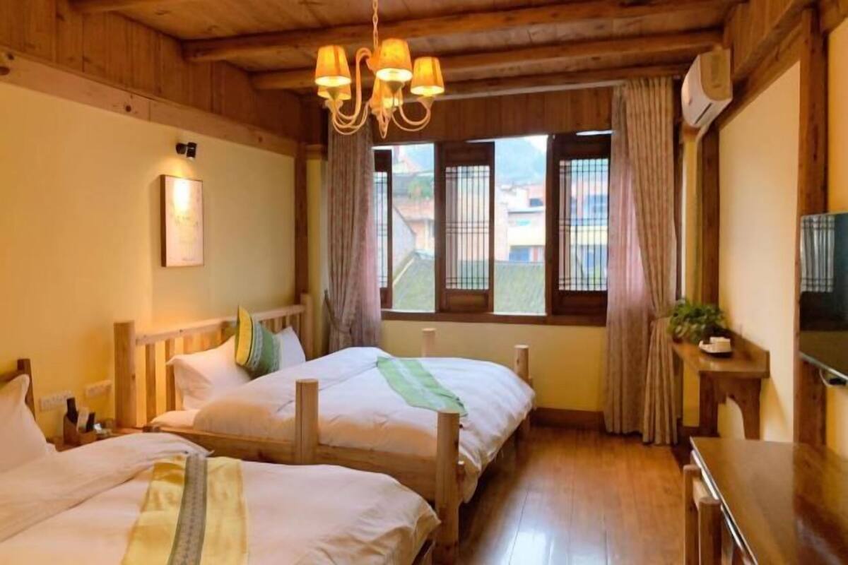 A room with two wooden beds, a chandelier, a window with curtains, and a small table with a plant.