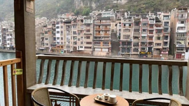 A balcony with wicker chairs and a small round table, overlooking a river with multi-story buildings in the background. - Mengqi Xiaozhu Inn (Zhenyuan County)