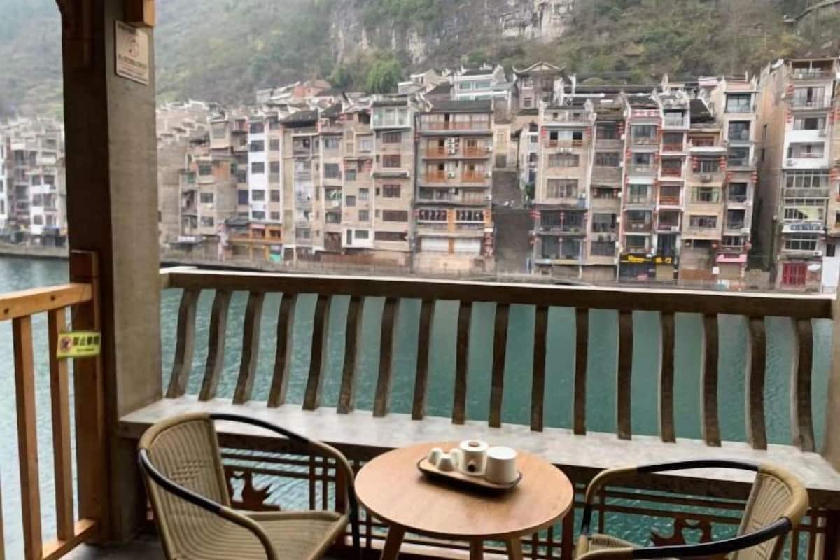 A balcony with wicker chairs and a small round table, overlooking a river with multi-story buildings in the background.