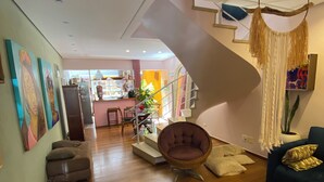 A spacious living area with a wooden floor, a round wicker chair, a dark blue sofa, and a staircase leading to an upper level.