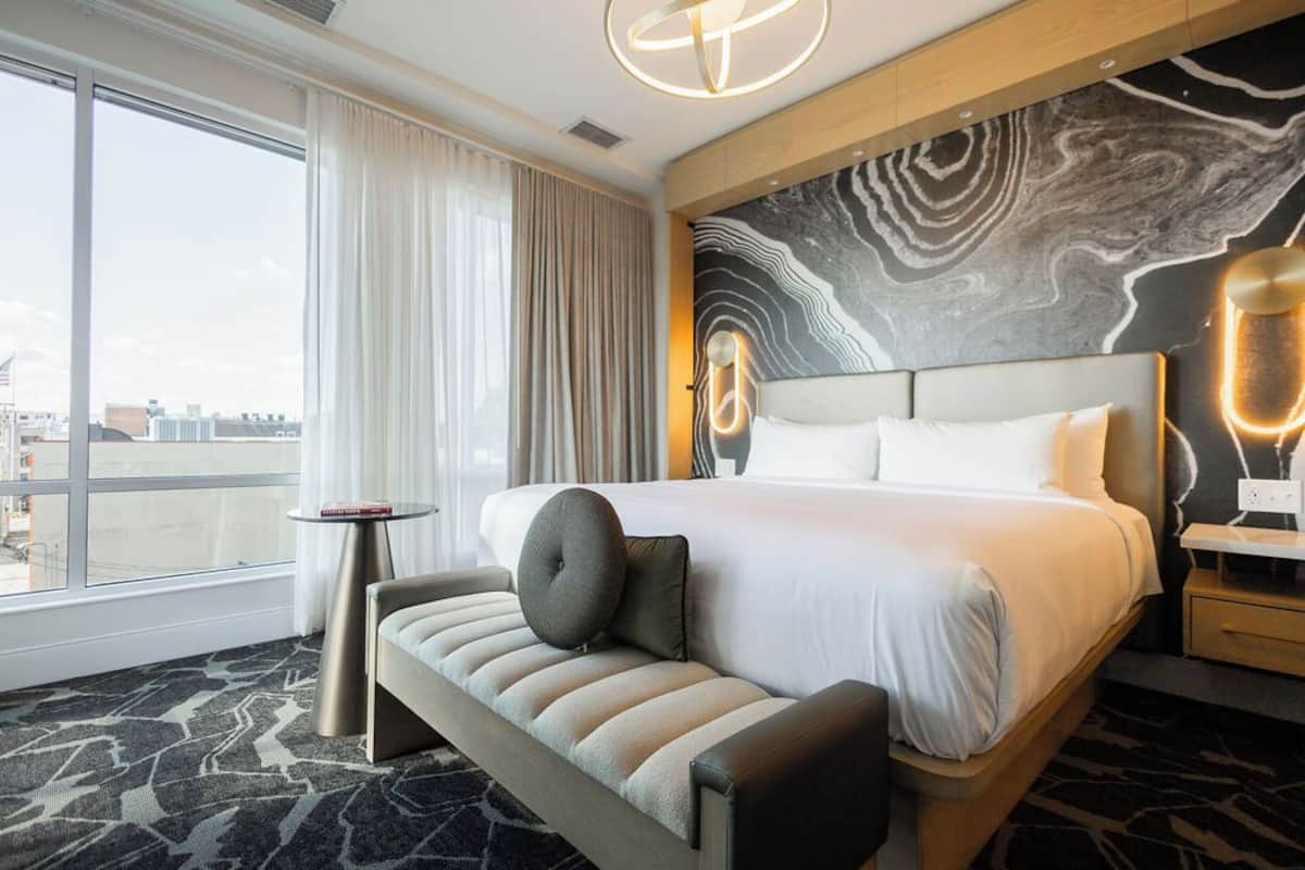 A modern hotel room with a large bed, a bench, a small table, and a city view.