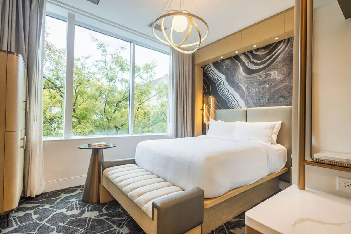 A modern hotel room with a large bed, a small round table, a bench, and a view of trees through large windows.