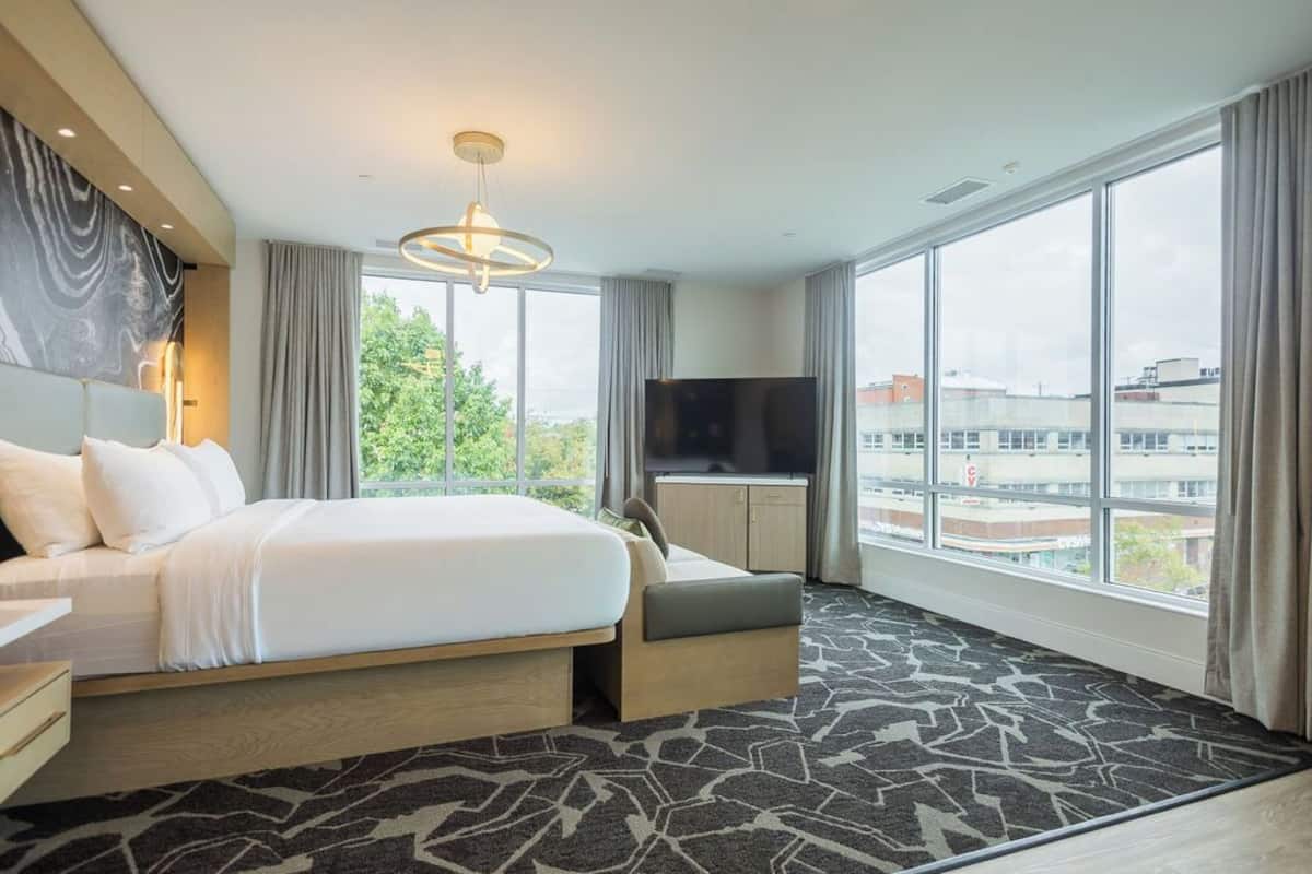 A modern hotel room with a large bed, a flat-screen TV, and a view of the city through large windows.