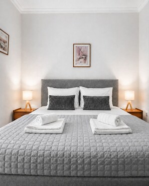 A neatly made bed with a grey quilt, two pillows, and two folded towels on the bed. There are two bedside tables with lamps, and a framed picture on the wall above the bed.