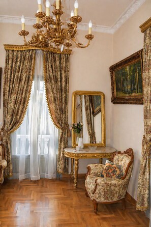 A room with a chandelier, ornate curtains, a mirror, a painting, and a floral-patterned chair.