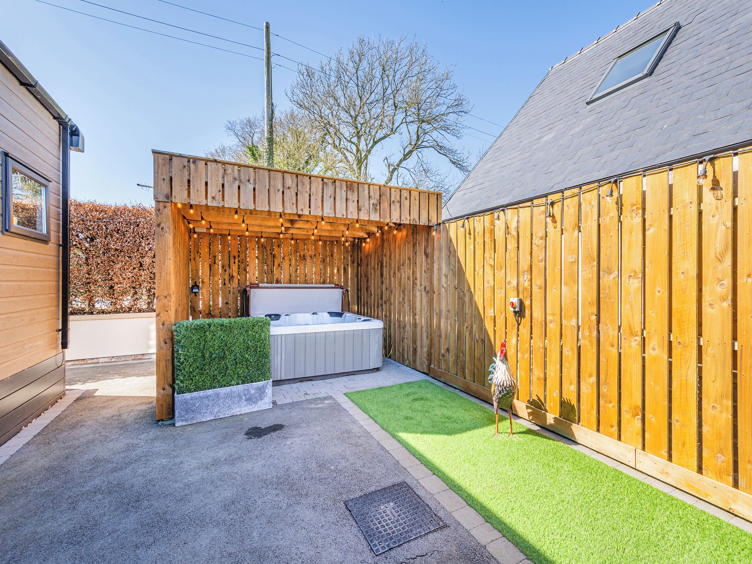 Un bain à remous installé dans une structure en bois, avec vue sur une haie et un bâtiment.