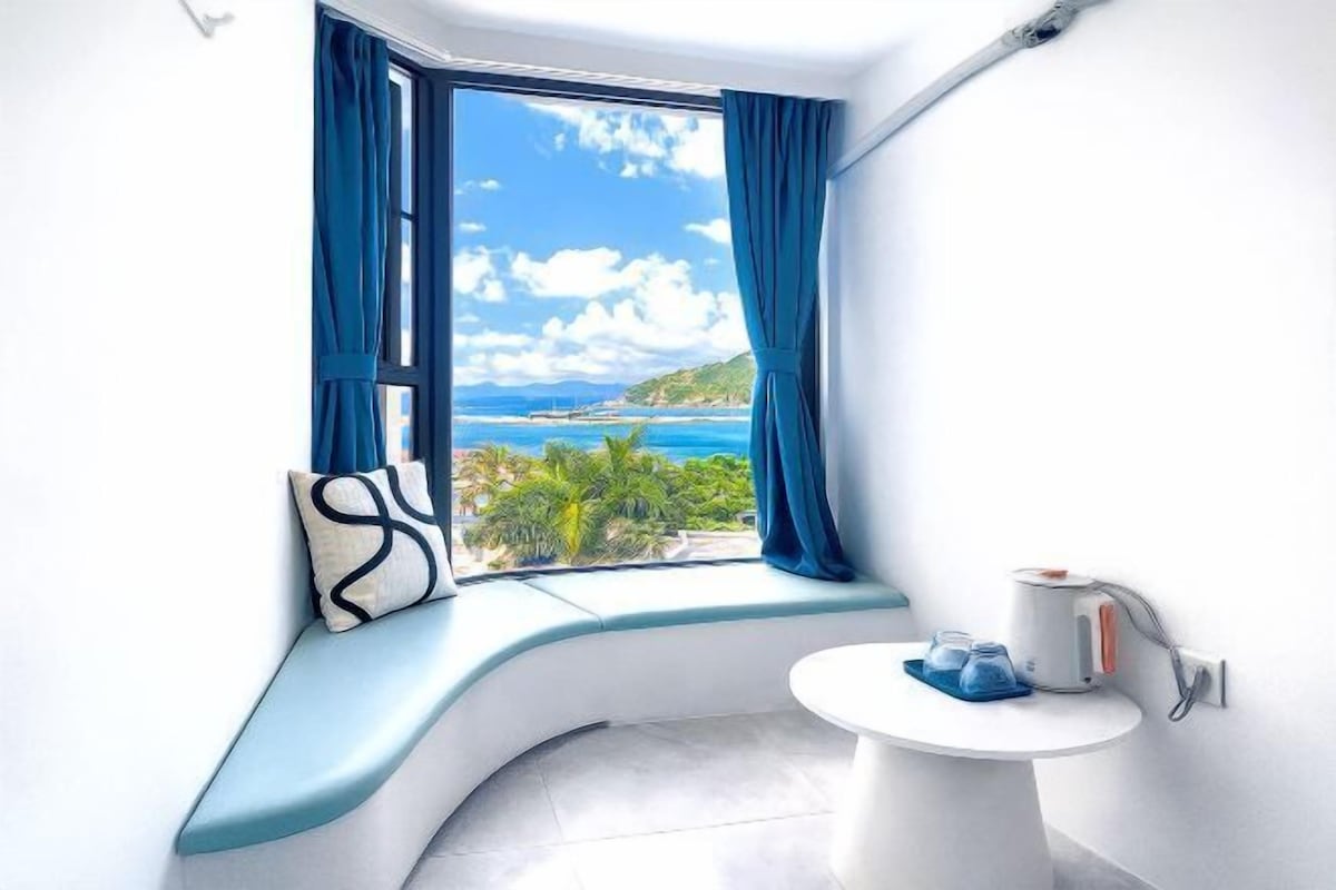 A bathroom with a curved bench, a small sink, and a window with a view of the sea.