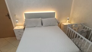 A bedroom with a large bed, two bedside tables with lamps, a white crib, and a tiled floor. - Modern apartment near beaches and airport (Tetuán)
