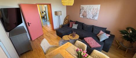 Een moderne woonkamer met een donkergrijze bank, een houten eettafel met een vaas roze bloemen, en een hanglamp.