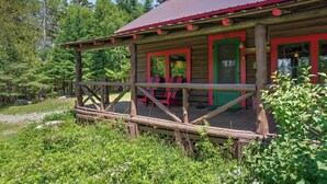 A rustic cabin with a wooden deck and a red roof, surrounded by lush greenery. - Vintage Log Cabin in the Heart of the Adirondacks (Indian Lake)