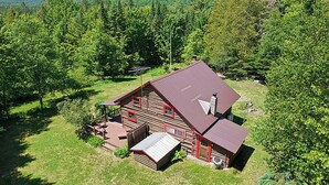 A log cabin with a metal roof, surrounded by dense forest and hills. - Vintage Log Cabin in the Heart of the Adirondacks (Indian Lake)