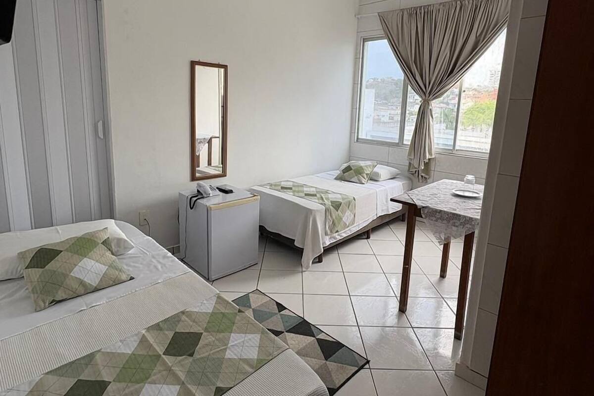 Habitación de hotel con dos camas, un escritorio, una silla, un ventilador de techo, una ventana con cortinas y vistas a la ciudad.