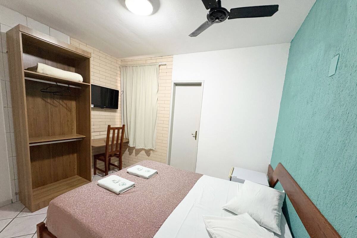 Un dormitorio con cama, un armario de madera, una silla, un escritorio, un ventilador de techo y una puerta.