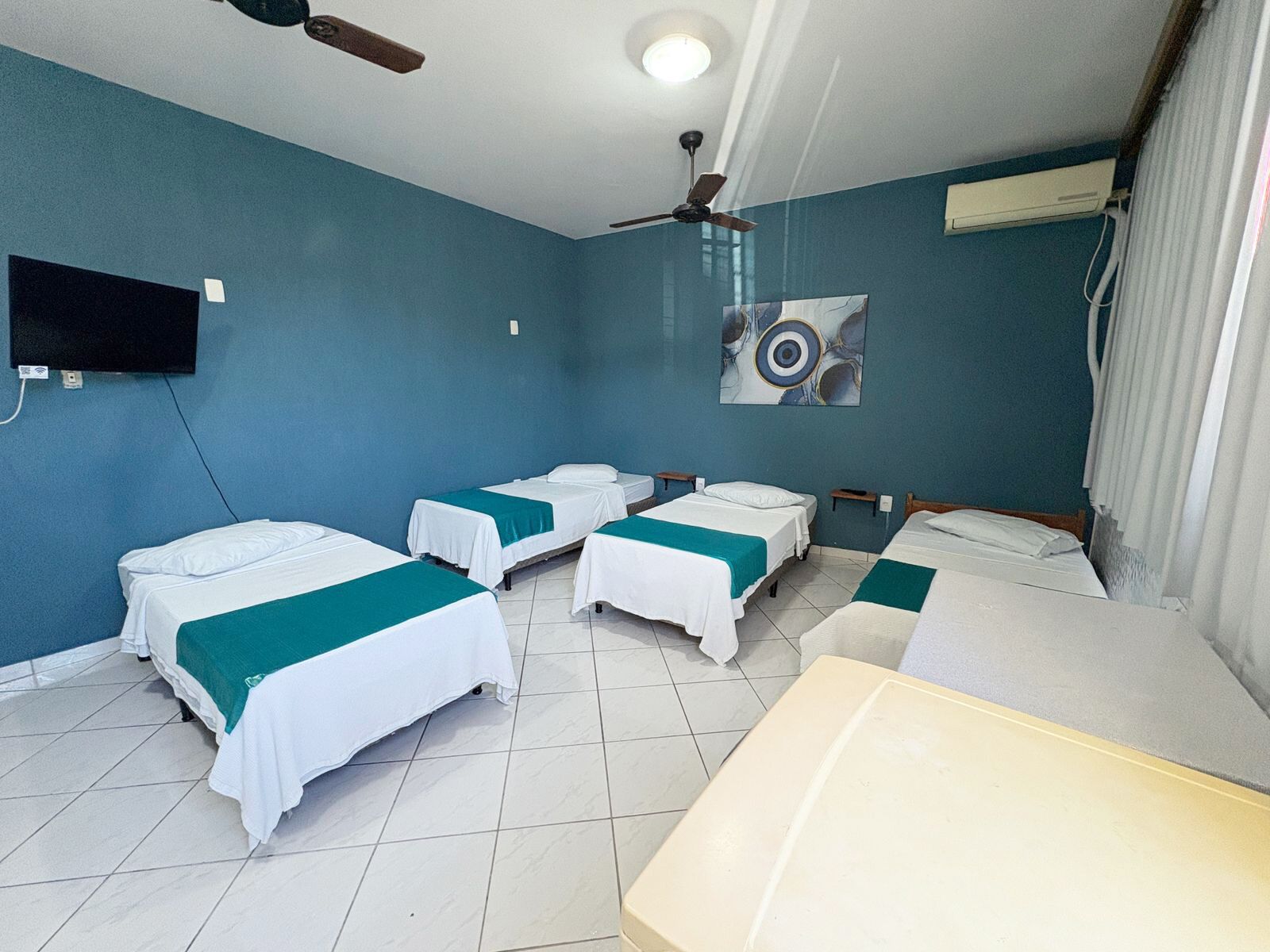 A hotel room with four single beds, a ceiling fan, a mounted TV, and a painting on the wall.