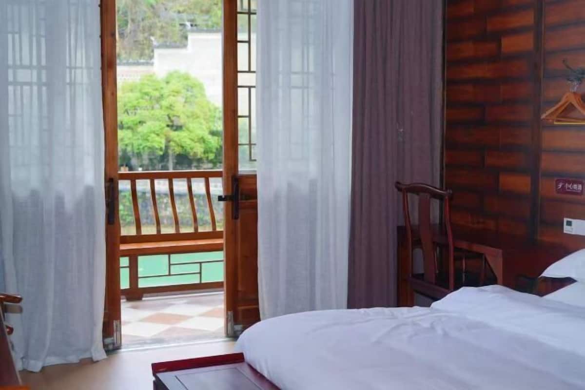 A hotel room with a bed, a wooden chair, a window with a view of greenery, and a brick wall.