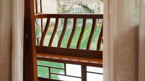 Une vue à travers une fenêtre ornée d’une balustrade en bois, donnant sur un espace extérieur verdoyant et sur des bâtiments en arrière-plan.