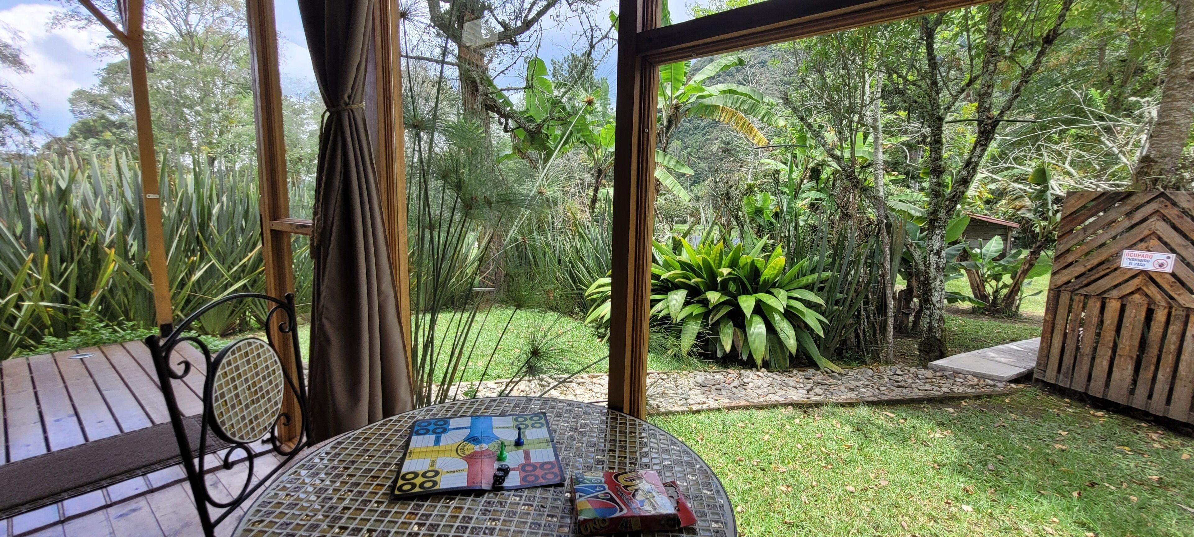 A garden view from a room with a table set for a game, surrounded by lush greenery and a wooden structure.