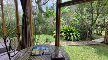 A garden view from a room with a table set for a game, surrounded by lush greenery and a wooden structure.