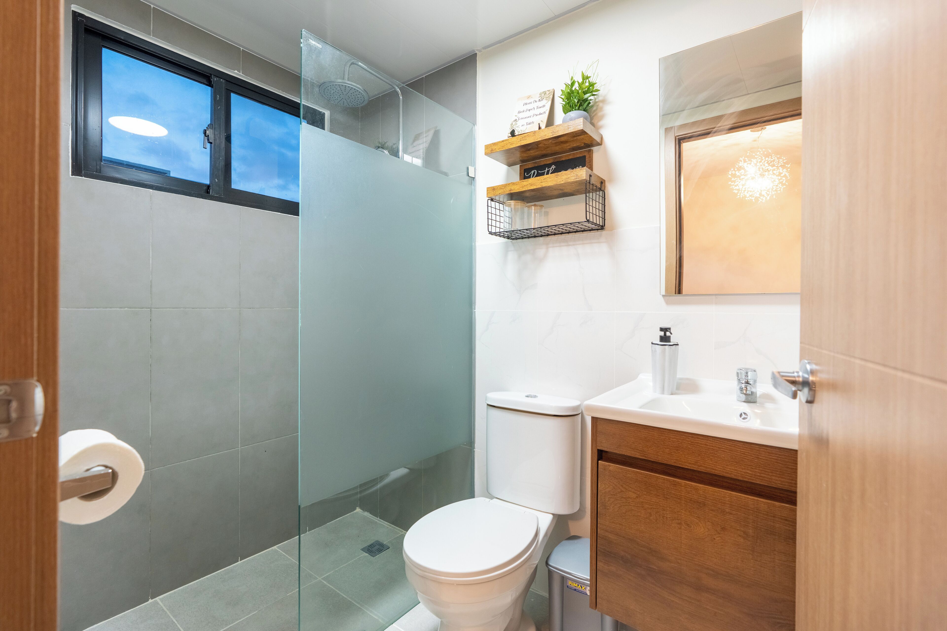 Un baño moderno con cabina de ducha de vidrio, inodoro, lavabo con espejo y estantes de madera con objetos decorativos.