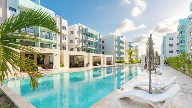 A swimming pool with lounge chairs and umbrellas, surrounded by multi-story buildings with balconies and outdoor seating areas.