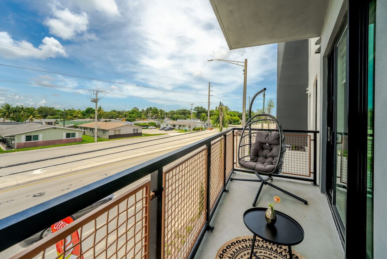 A balcony with a hanging chair, a small table with a vase, and a view of a road and houses.