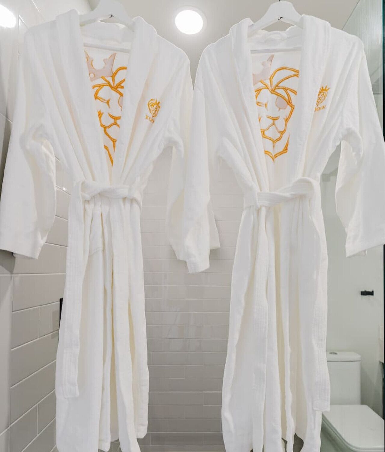 Two white bathrobes with gold embroidery hanging in a bathroom with white tiles and a white toilet.