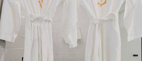 Two white bathrobes with gold embroidery hanging in a bathroom with white tiles and a white toilet.