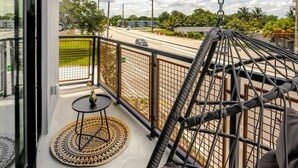 A balcony with a hanging chair, a small round table with a vase, and a view of a road and greenery. - Lux Design Apartment near beach (Fort Lauderdale)