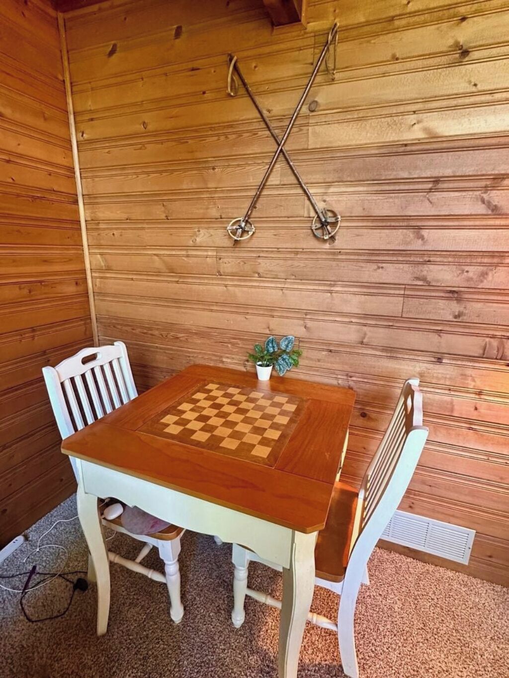 A wooden cabin with a checkered table and two chairs, a small potted plant, and a metal cross hanging on the wall.